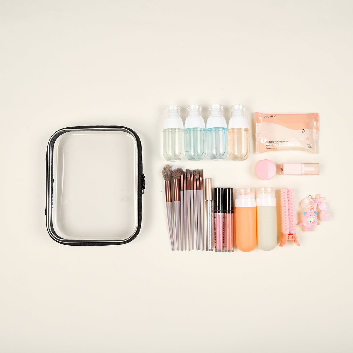 A clear zipper case with black trim beside travel bottles, makeup brushes, lip glosses, a compact, a pack of wet wipes and a charm, laid out on a cream background