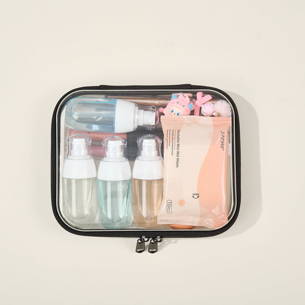 A clear zipper case with black trim holding three pump bottles, a spray bottle, makeup brushes, a charm and a pack of wet wipes, on a cream background